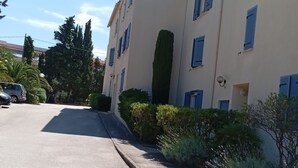 Un immeuble à plusieurs étages, doté d’une façade beige et de volets bleus, d’une allée pavée et entouré de verdure.
