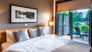 Un dormitorio moderno con una cama grande, un cuadro enmarcado en la pared, una lámpara de noche y una puerta corrediza de vidrio que da a un espacio exterior con sillas.