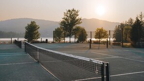 An outdoor tennis court with a net, surrounded by trees and a view of a body of water and hills in the background. - Lakefront & Mountain View - 1 Bedroom Resort Suite (Lac-Supérieur)