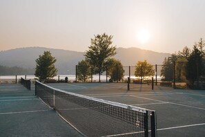 An outdoor tennis court with a net, surrounded by trees and a view of a body of water and hills in the background.