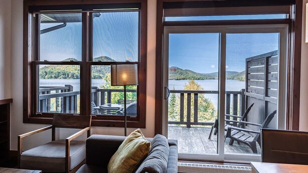 Sala de estar com sofá, cadeiras e mesa. Janelas amplas com vista para um deck e um corpo de água.