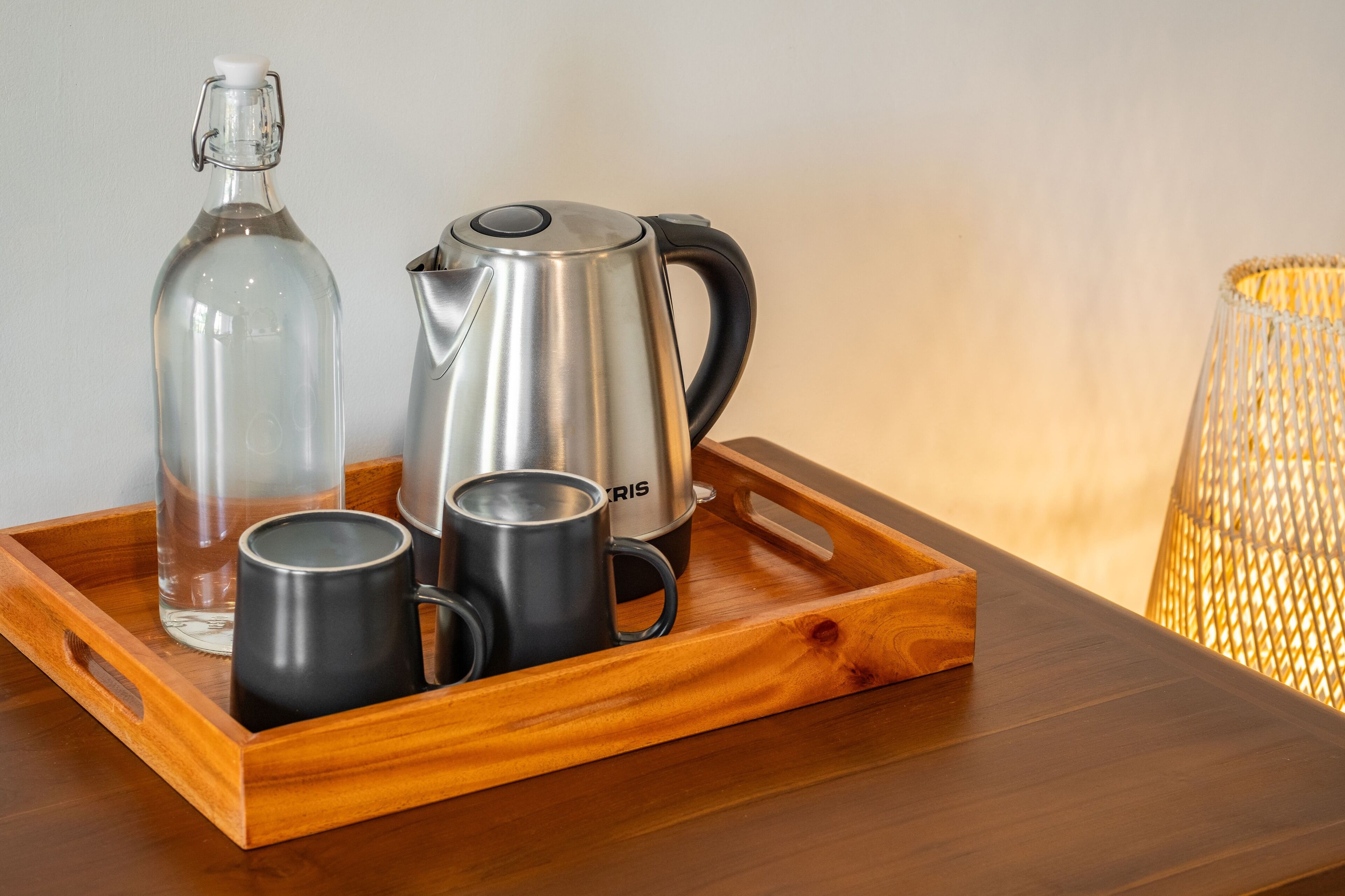 A wooden tray with a glass bottle, a stainless steel kettle, and two black mugs on a wooden table.