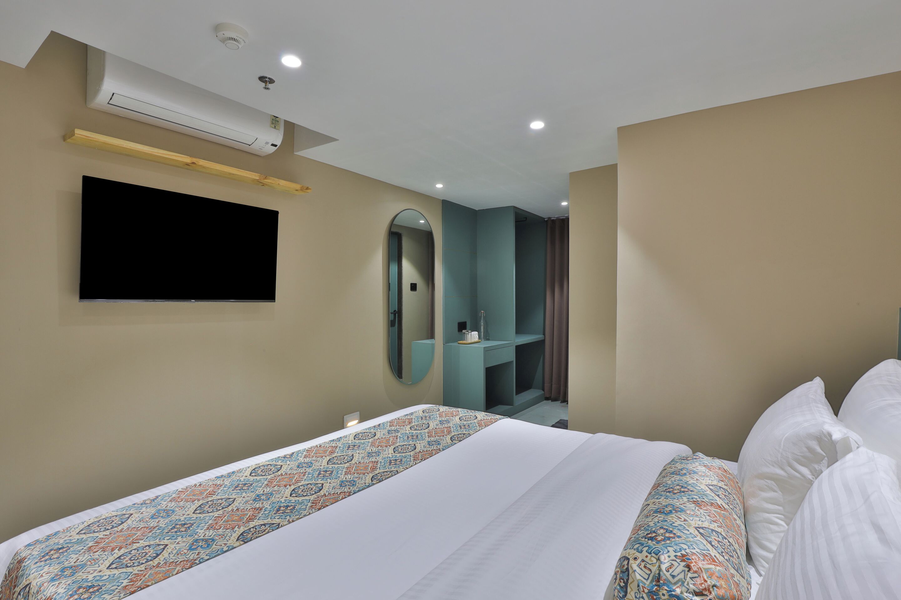 A hotel room with a bed, a flat-screen TV mounted on the wall, a mirror, and a small vanity area with a sink.