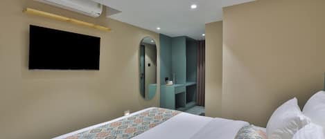 A hotel room with a bed, a flat-screen TV mounted on the wall, a mirror, and a small vanity area with a sink.