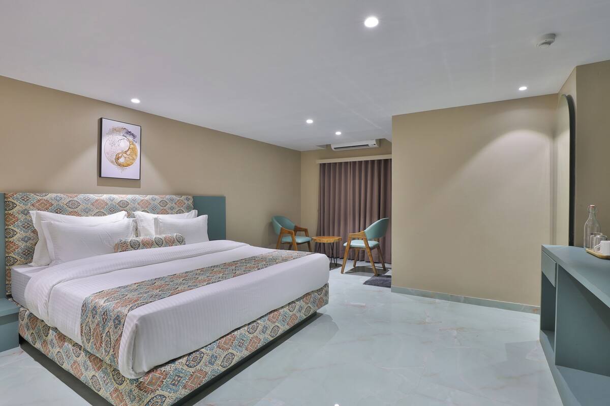 A modern hotel room with a large bed, a small seating area with chairs, and a desk with a bottle and glasses.