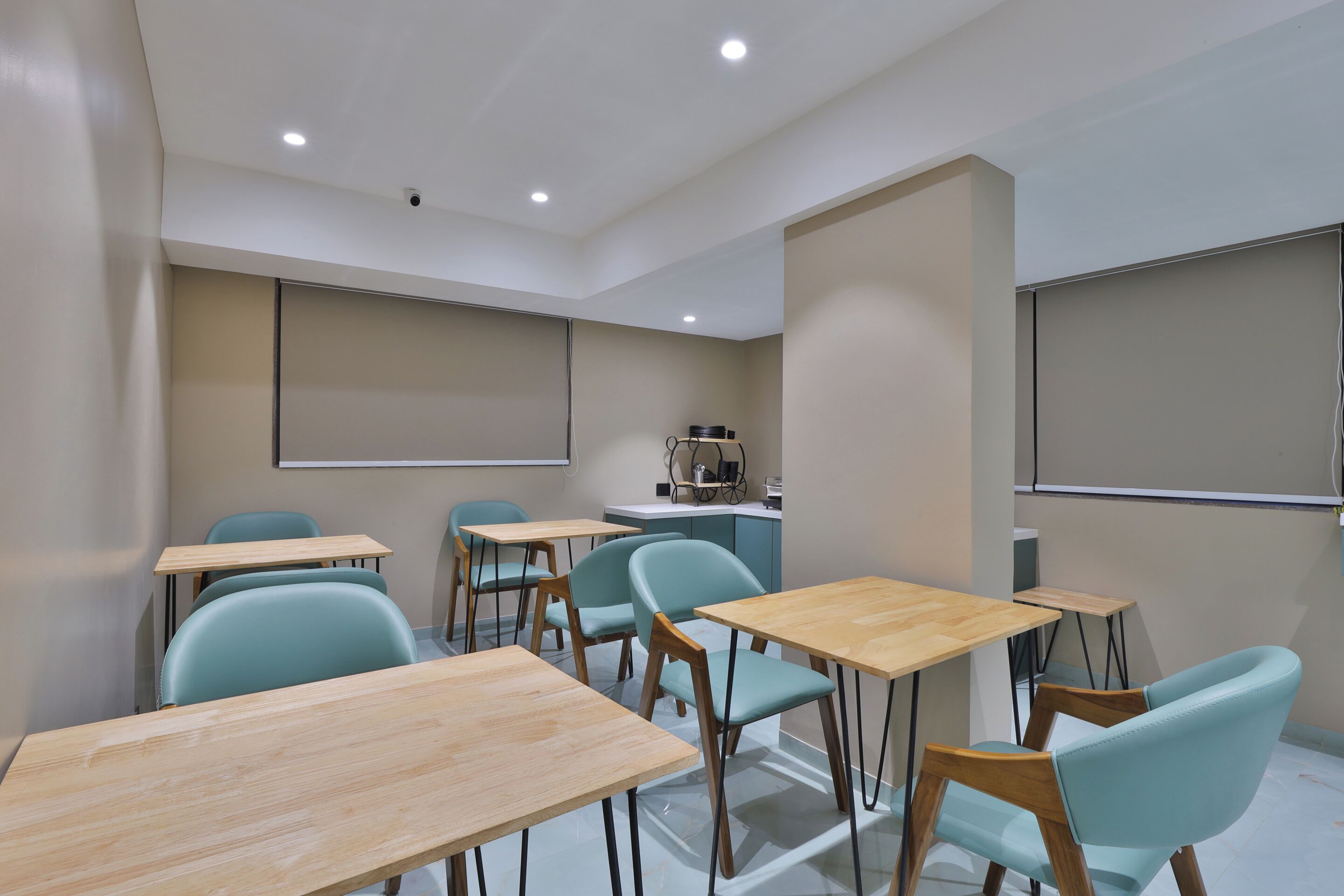 A classroom with wooden desks and teal chairs, a projector screen, and a kitchenette area.