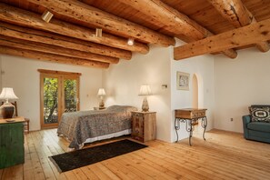 Un dormitorio con techo de madera, una cama con colcha estampada, dos lámparas de noche, una mesita de noche de madera, una pequeña mesa con lámpara, una silla verde y vista al exterior a través de una ventana.