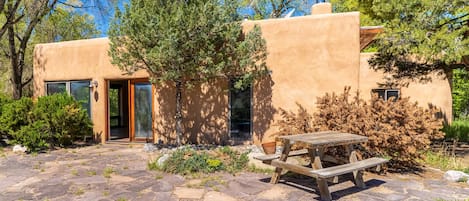 Un edificio con un patio empedrado de piedra, una mesa de picnic de madera y un árbol grande al frente.