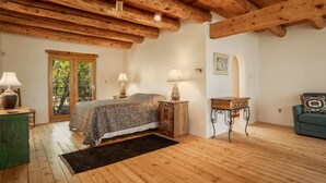 Ett sovrum med ett träpanelat tak, en säng med blommigt sänglinne, två sänglampor, ett träbord som sängbord, ett litet bord med en lampa, en grön soffa och ett fönster med utsikt över träd.