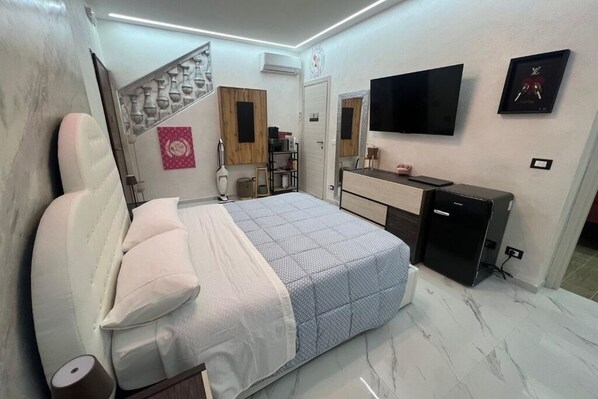 Un dormitorio moderno con una cama, televisor, espejo y un refrigerador pequeño.