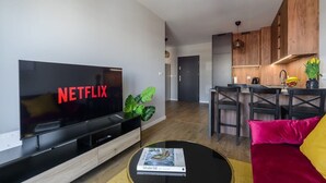 A modern living room with a flat-screen TV displaying 'NETFLIX', a dining area with a kitchen in the background, and a small table with a magazine. - Spacious apartment on Wróblewskiego (Łódź)