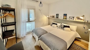 A bedroom with a bed, a hanging light fixture, a shelf with pictures, a wardrobe, and a window with curtains.