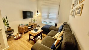 A living room with a sofa, a coffee table, a TV, a plant, and a window with blinds.