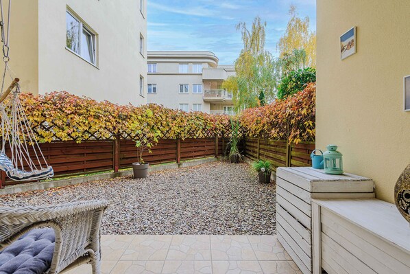 A courtyard with a wicker chair, a wooden bench, a small table with a lantern, and a wooden fence covered in autumn leaves.