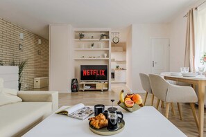 A modern living room with a flat-screen TV displaying "NETFLIX," a dining area with a wooden table and chairs, and a cozy seating area with a sofa and a coffee table with breakfast items.