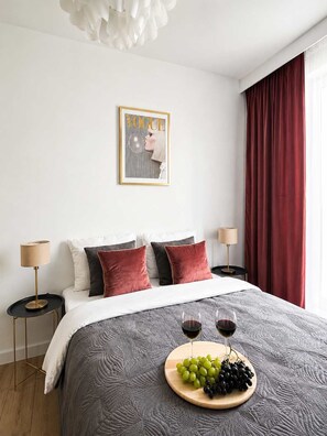 Een netjes opgemaakt slaapkamer met een bed, twee bedlampjes, een lijst met een afbeelding aan de muur en een raam met rode gordijnen.