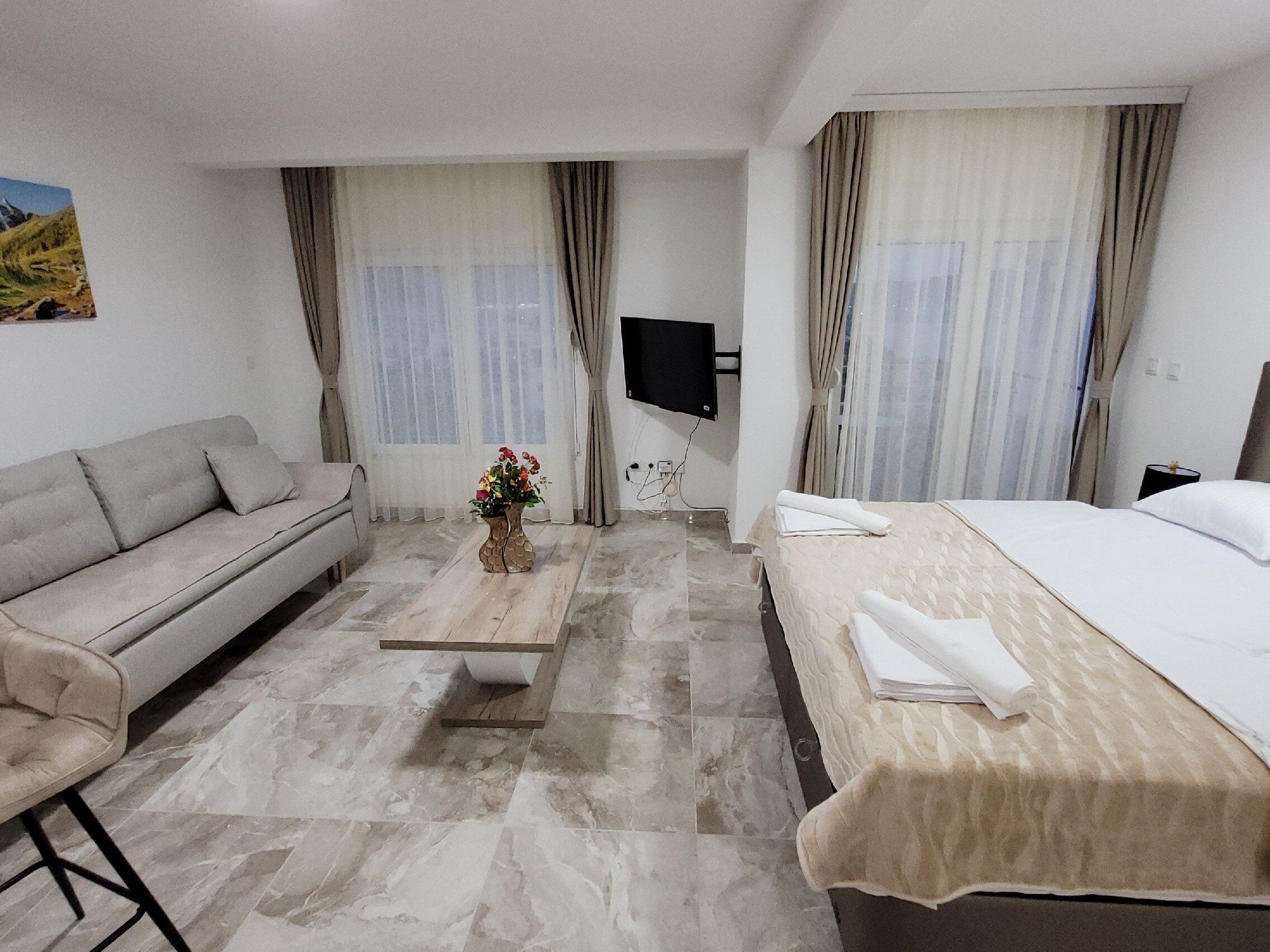 A hotel room with a sofa, a bed, a wooden coffee table with a vase, and a TV mounted on the wall.