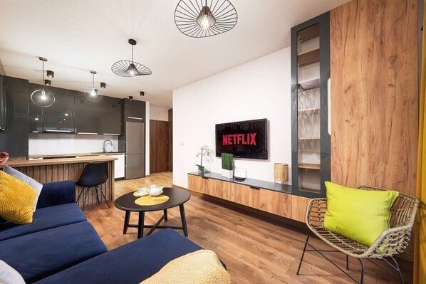 A modern living room with a sofa, a coffee table, and a wicker chair. A flat-screen TV is mounted on the wall, and there is a kitchen area in the background.