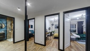 A modern hotel room with a wooden floor, a bed with a green blanket, a desk with a chair, and a bathroom with a shower and a mirror.