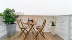 A wooden table with two chairs, a bottle of wine, and a fruit bowl on a balcony with plants and a view of buildings.