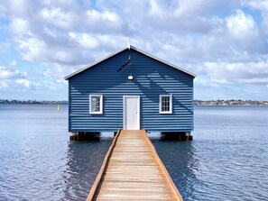 A blue wooden house with a white door and windows, connected to a wooden dock extending into calm water.