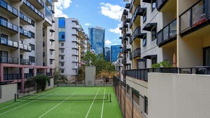 En tennisbana omgiven av bostadshus med balkonger, en klar himmel och en modern byggnad i bakgrunden.