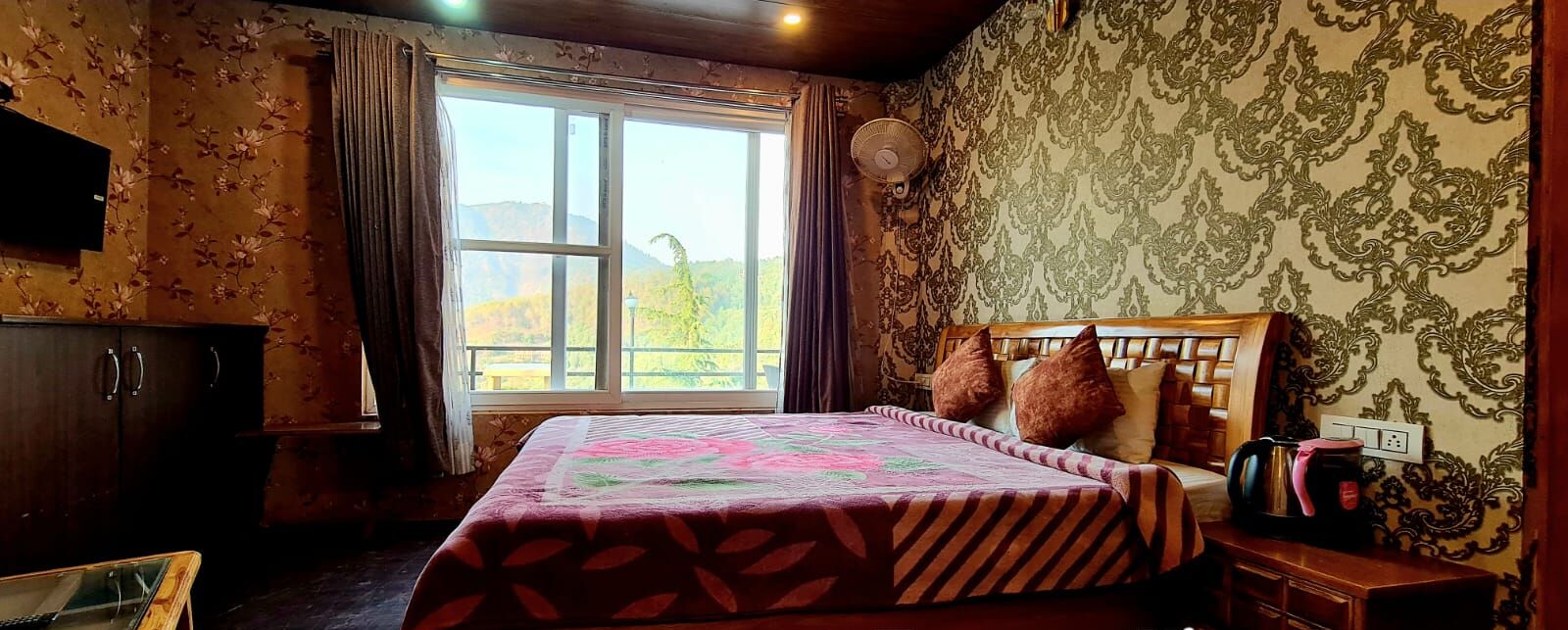 A bedroom with a large bed, a wooden headboard, a window with curtains, and a view of a mountainous landscape.