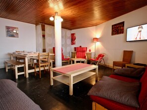 A room with a wooden ceiling, a dining area with wooden tables and chairs, a sofa, a television, and a red accent wall. - Appartement au cœur de Chamrousse, 50m² avec balcon, pied des pistes, animaux acceptés (Chamrousse)