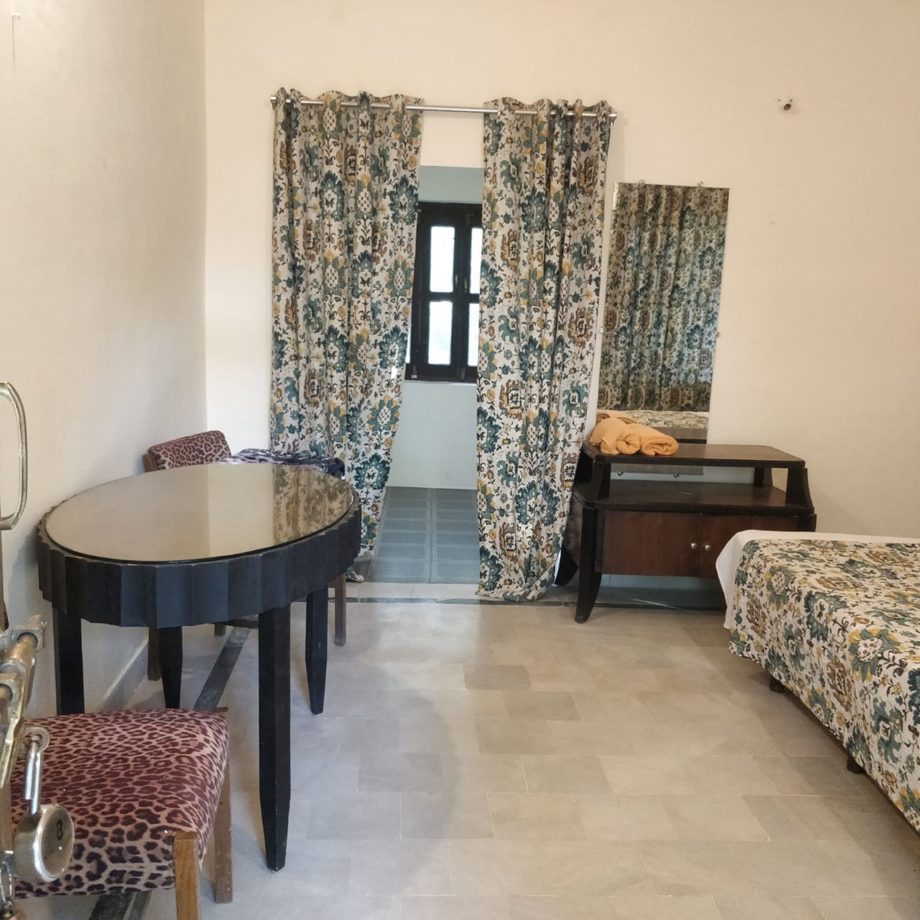 Una habitación pequeña y amoblada con una mesa redonda, dos sillas, una cama con colcha estampada y un aparador de madera.
