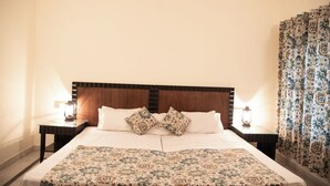 A neatly made bed with a patterned bedspread, two matching pillows, and a wooden headboard. There are two bedside tables with lamps, one on each side of the bed. The room has plain walls and a patterned curtain on the window. - Shivika Lake Hotel Ranakpur (Desuri)