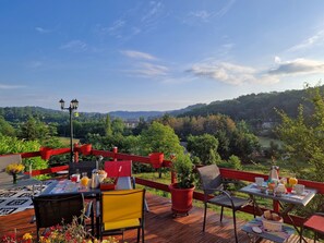 An outdoor dining area with tables and chairs, surrounded by a wooden deck and red railings, overlooking a lush valley and distant hills.
