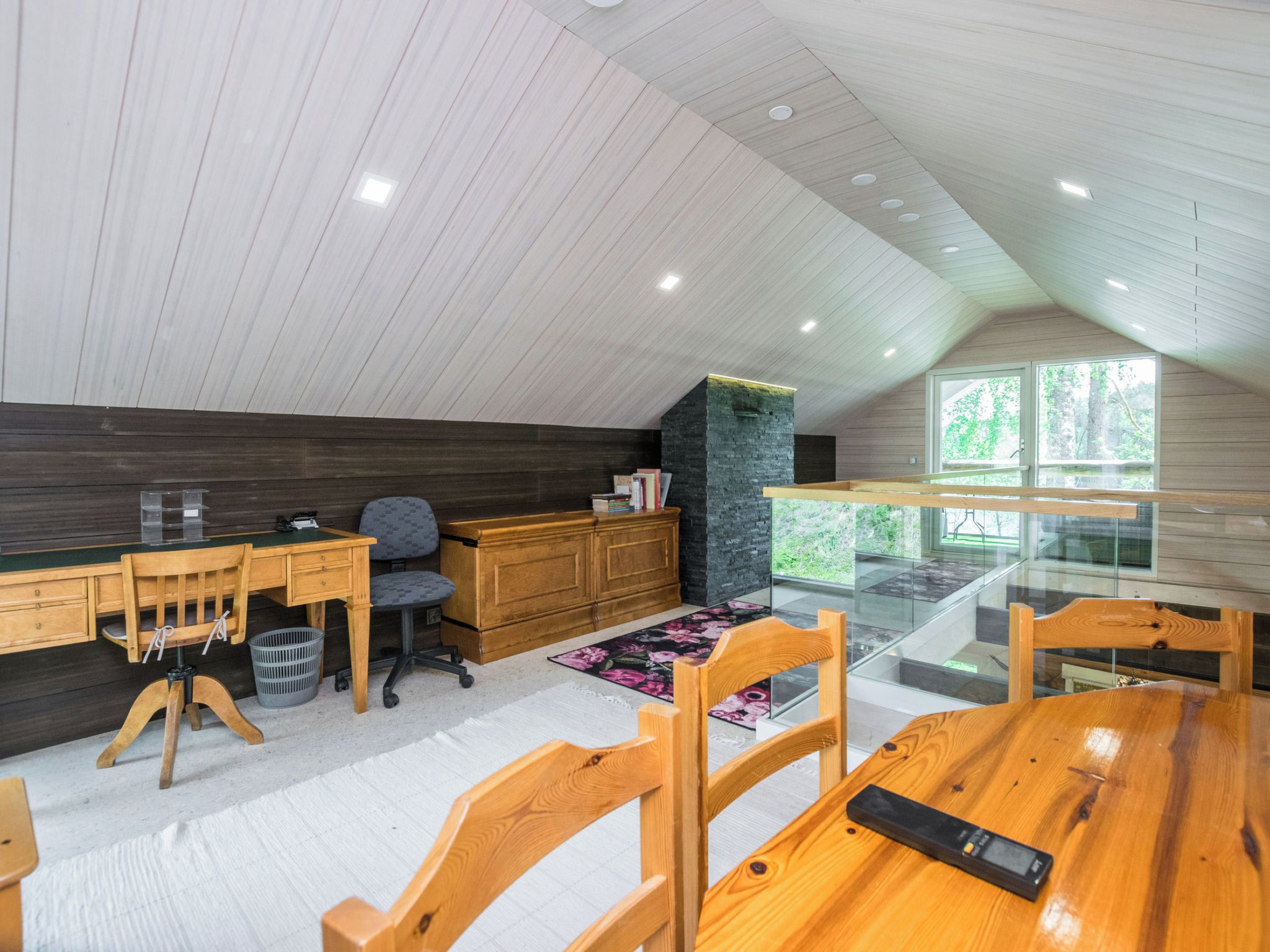 Ein modernes Schlafzimmer im Loft-Stil mit schrägem Dach, Holzmöbeln, einer Glasabtrennung und Blick ins Grüne.