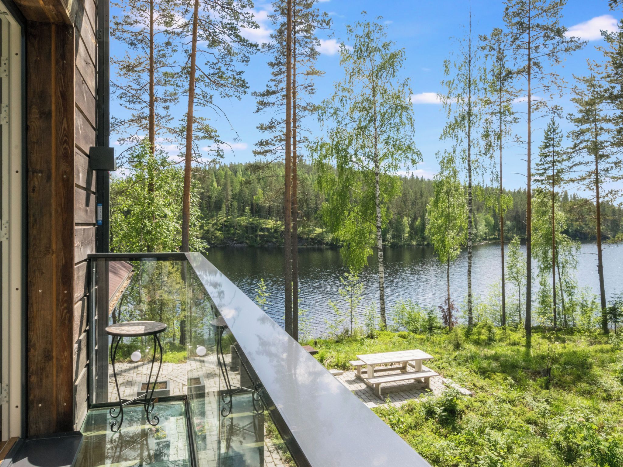 Parveke, jossa on lasikaide, ja josta avautuu näkymä järvelle. Parveketta ympäröivät puut ja sen vieressä on puinen rakennus.