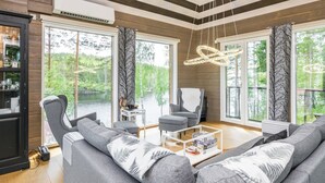 A modern living room with a large sofa, a chair, and a coffee table. There are large windows with curtains, a fireplace, and a ceiling with exposed beams.