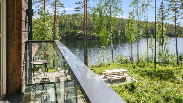 A balcony with glass railings overlooking a lake, surrounded by trees and a wooden building.