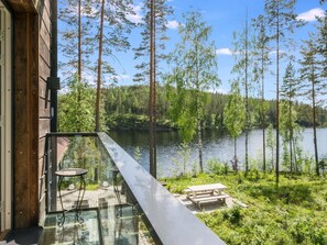 Un balcon doté de rampes en verre, donnant sur un lac, entouré d’arbres et bordé d’un bâtiment en bois.