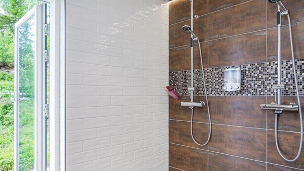 Un baño moderno con una puerta de cristal que da a un balcón, ducha con cabezal de lluvia y pared revestida con mosaicos.