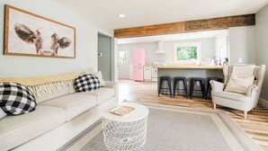 Una sala moderna con sofá, mesa de centro y un cuadro de vacas. Hay una zona de cocina con un refrigerador rosa y taburetes.