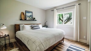 Un dormitorio con cama de madera, una mesita de noche con lámpara, una ventana con cortinas y un estante en la pared con objetos decorativos.
