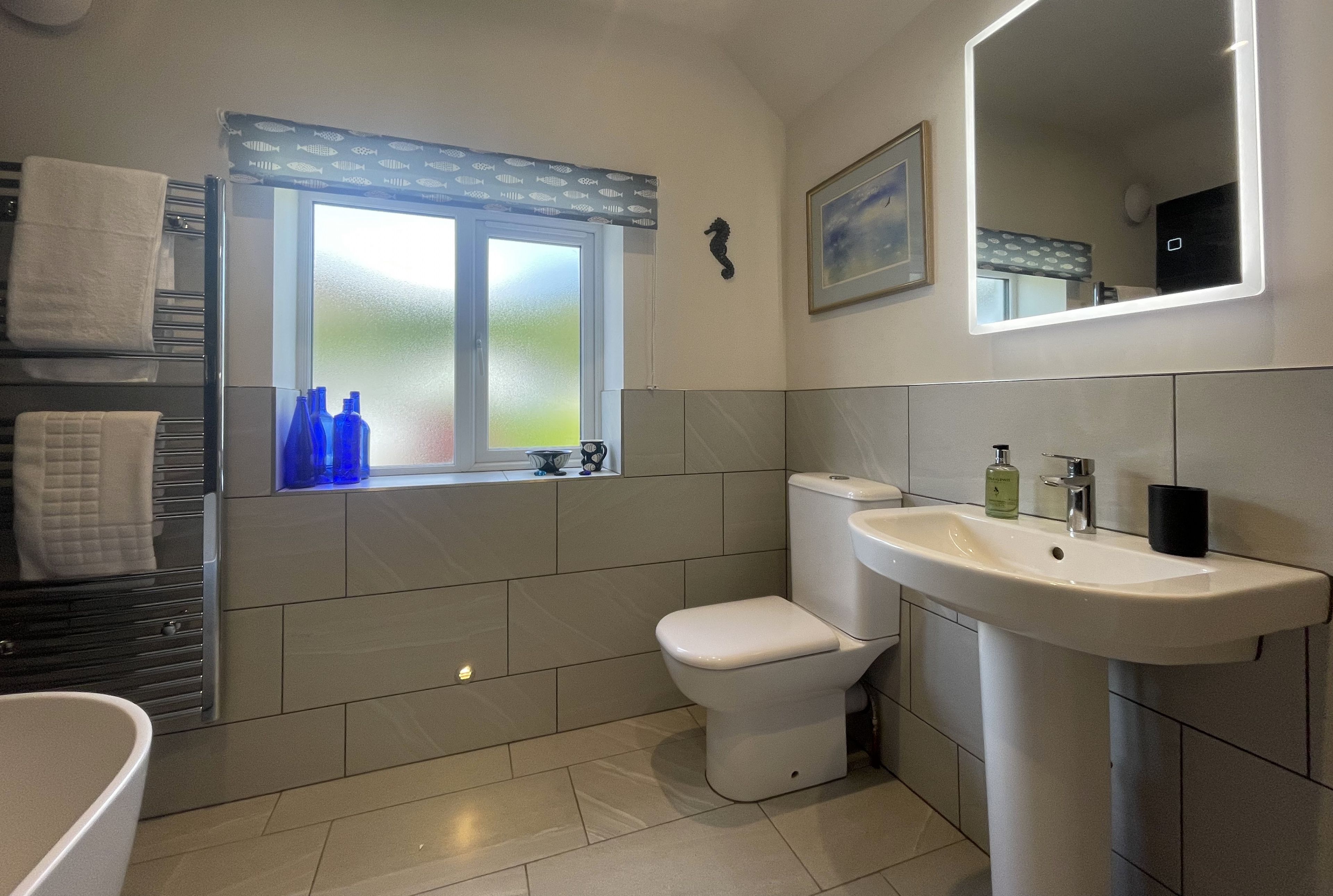 Een moderne badkamer met toilet, wastafel en ligbad. Er is een raam met geëtste glas en een handdoekrek met handdoeken.