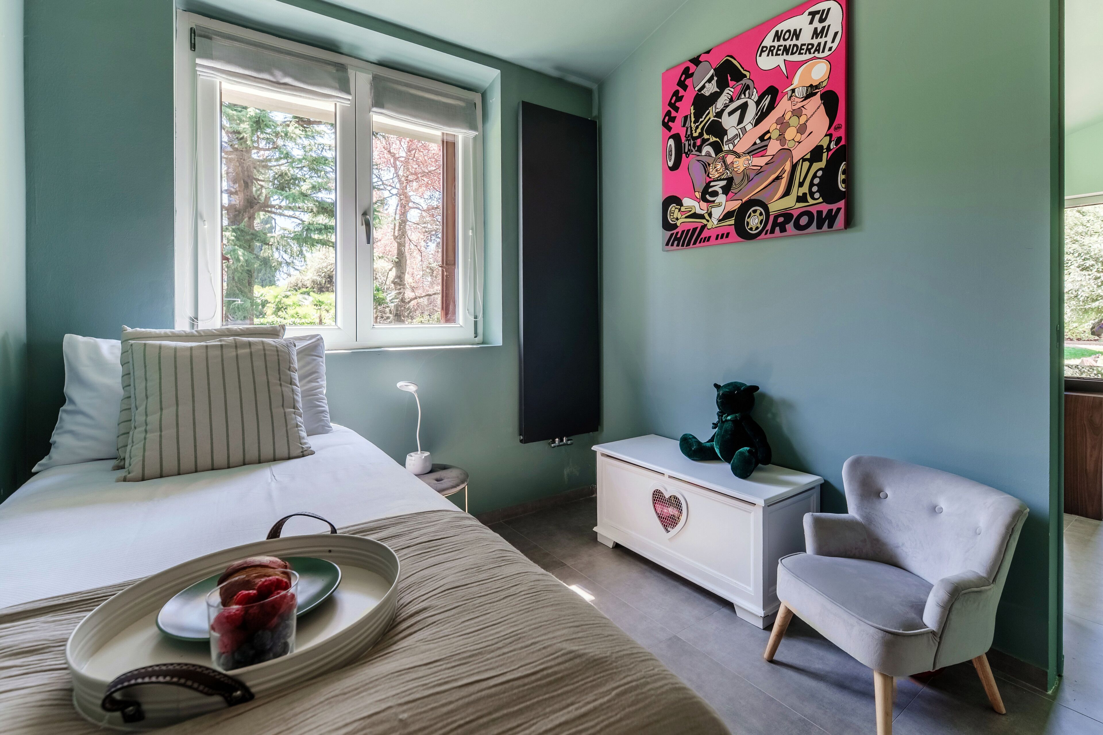 A bedroom with a bed, bedside table, dresser, chair, and a window with a view of trees.