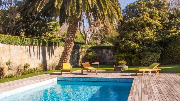 A rectangular swimming pool with a wooden deck surrounding it, featuring two lounge chairs and a palm tree. - Liiiving in Porto Garden Pool House (Porto)