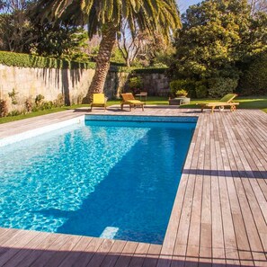 A rectangular swimming pool with a wooden deck surrounding it, featuring two lounge chairs and a palm tree. - Liiiving in Porto Garden Pool House (Porto)