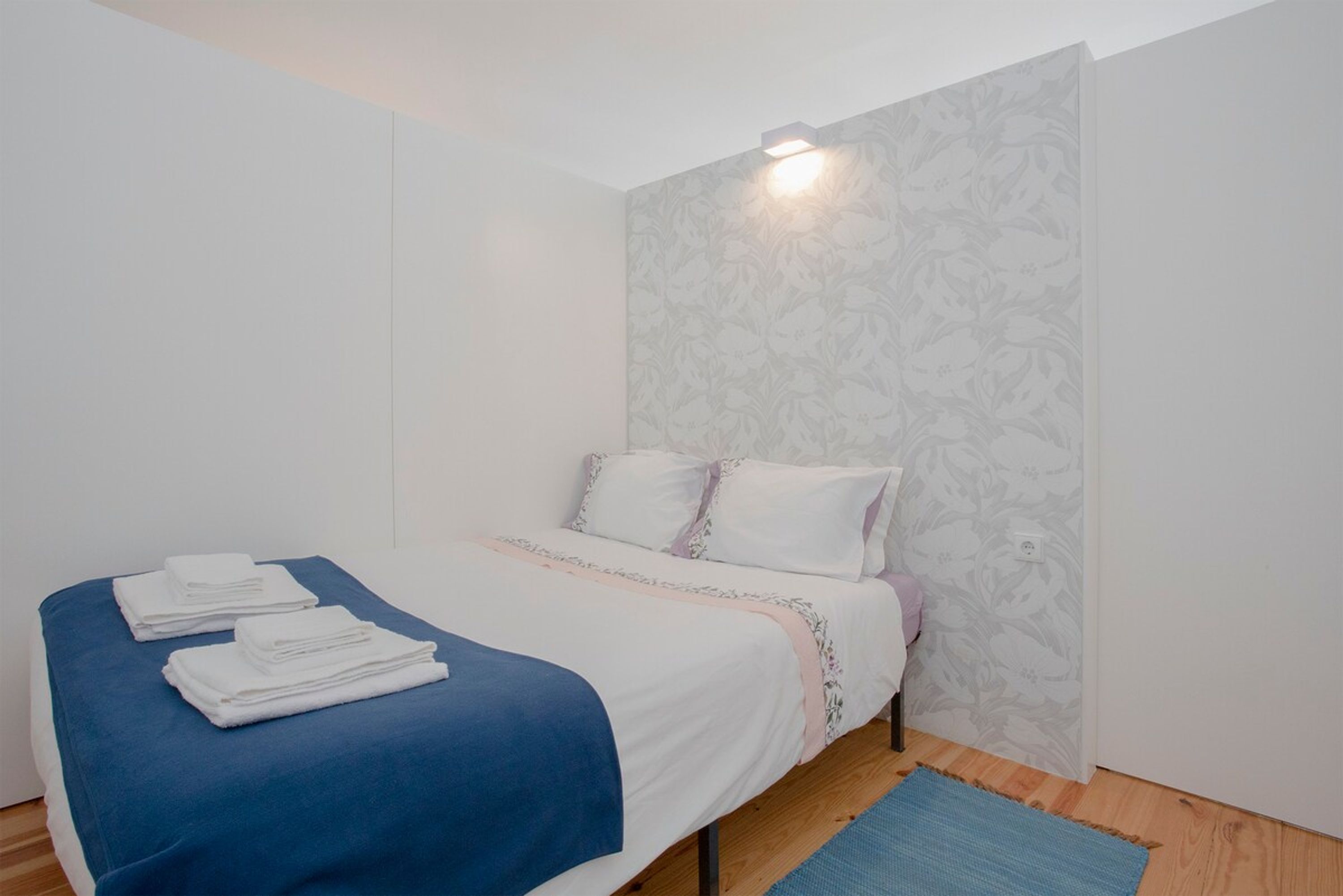 A neatly made bed with white and blue bedding, a floral-patterned wall, and a small light fixture above the bed.