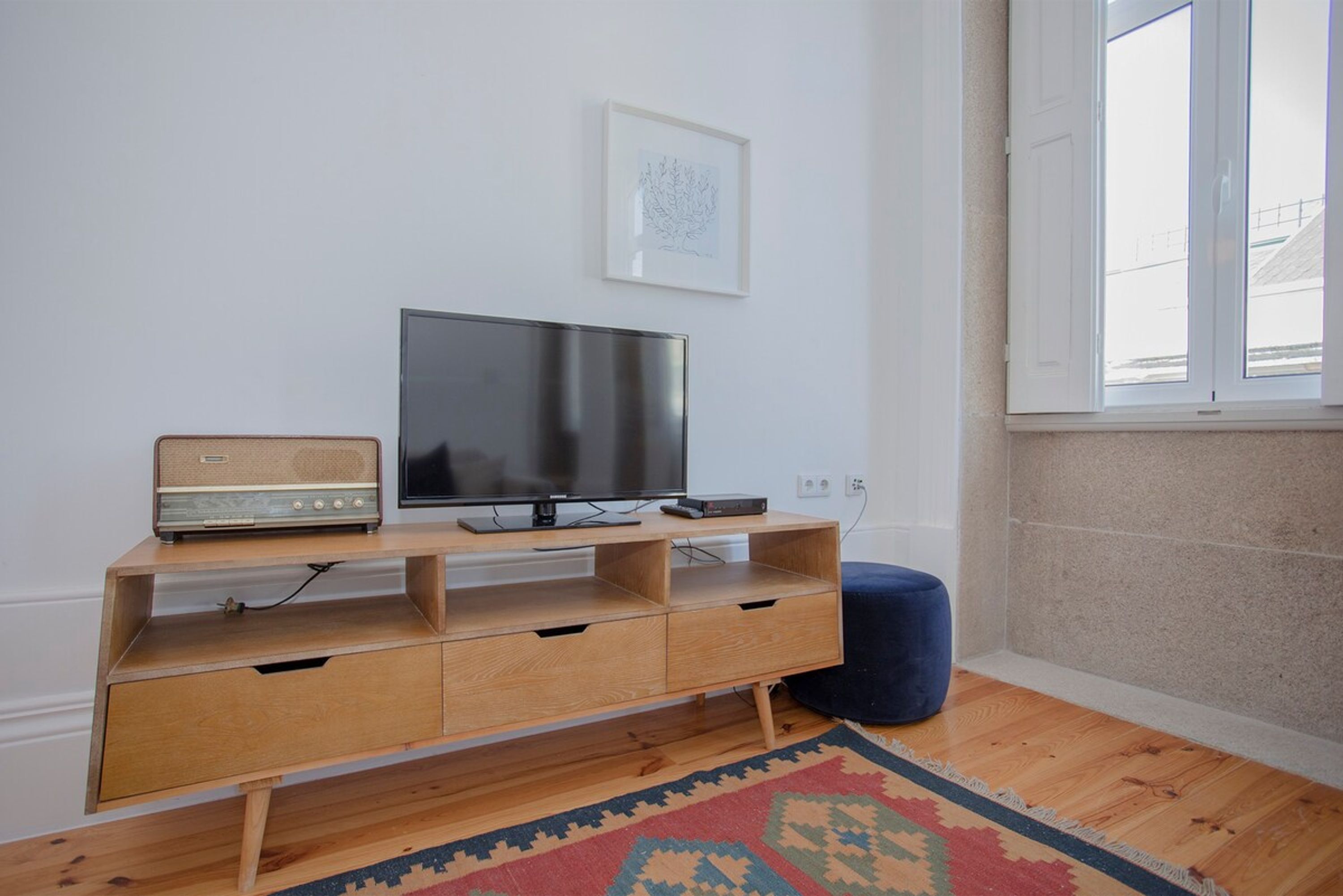 A modern living room with a wooden TV stand, a flat-screen TV, a vintage radio, and a blue ottoman.