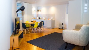 Une cuisine moderne avec des meubles blancs, un coin repas avec une table en bois et des chaises colorées, et un fauteuil gris dans un salon agrémenté d’un tapis foncé.