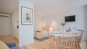 Une pièce moderne et bien éclairée, comprenant un lit, une table à manger avec des chaises, un téléviseur fixé au mur et un tableau encadré accroché au mur.
