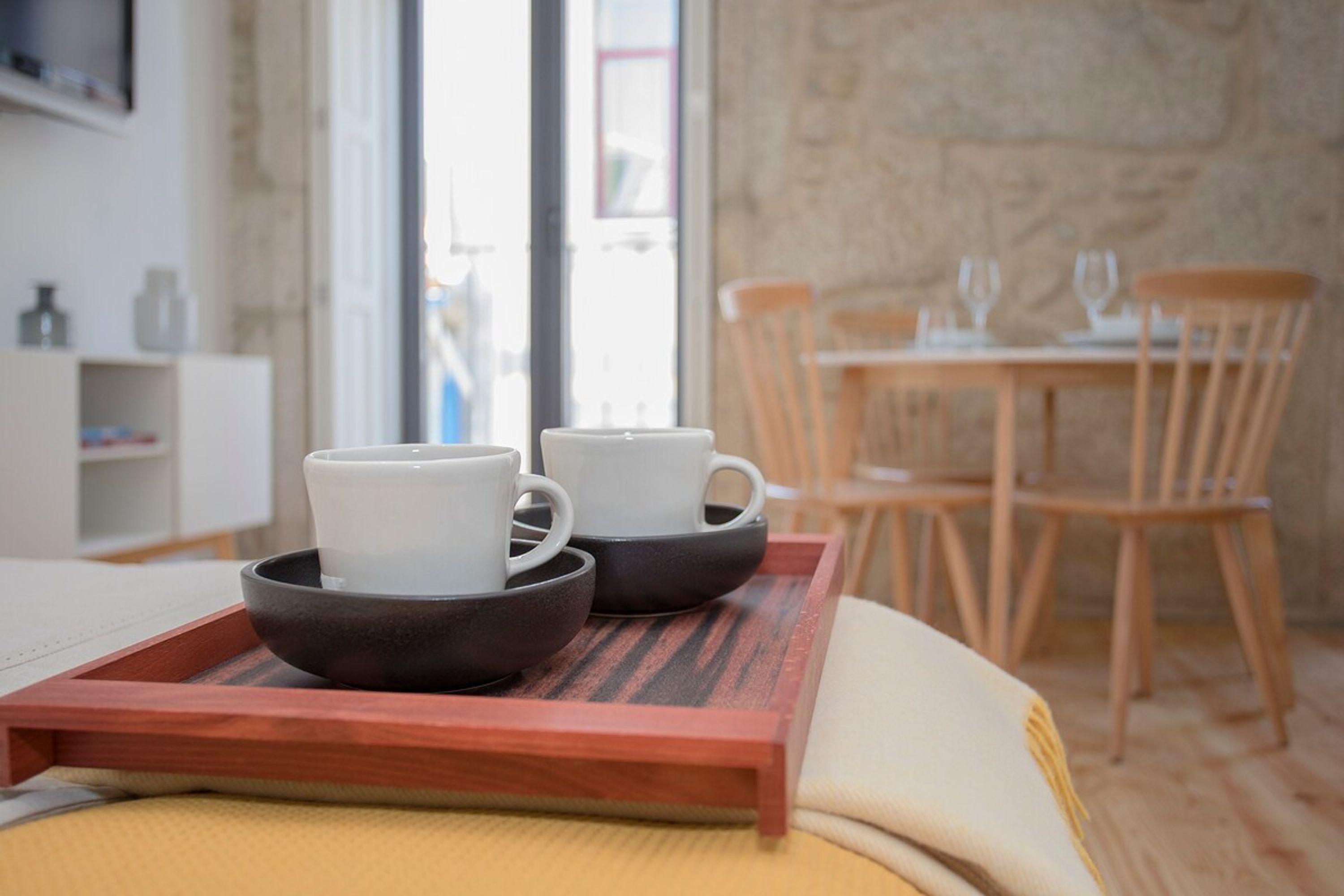 A wooden tray with two white cups and two black bowls on a bed.