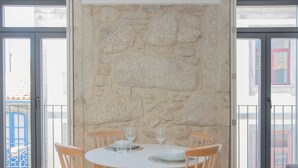 Een eethoek met een ronde tafel en vier houten stoelen, een stenen muur en grote ramen met uitzicht op gebouwen.