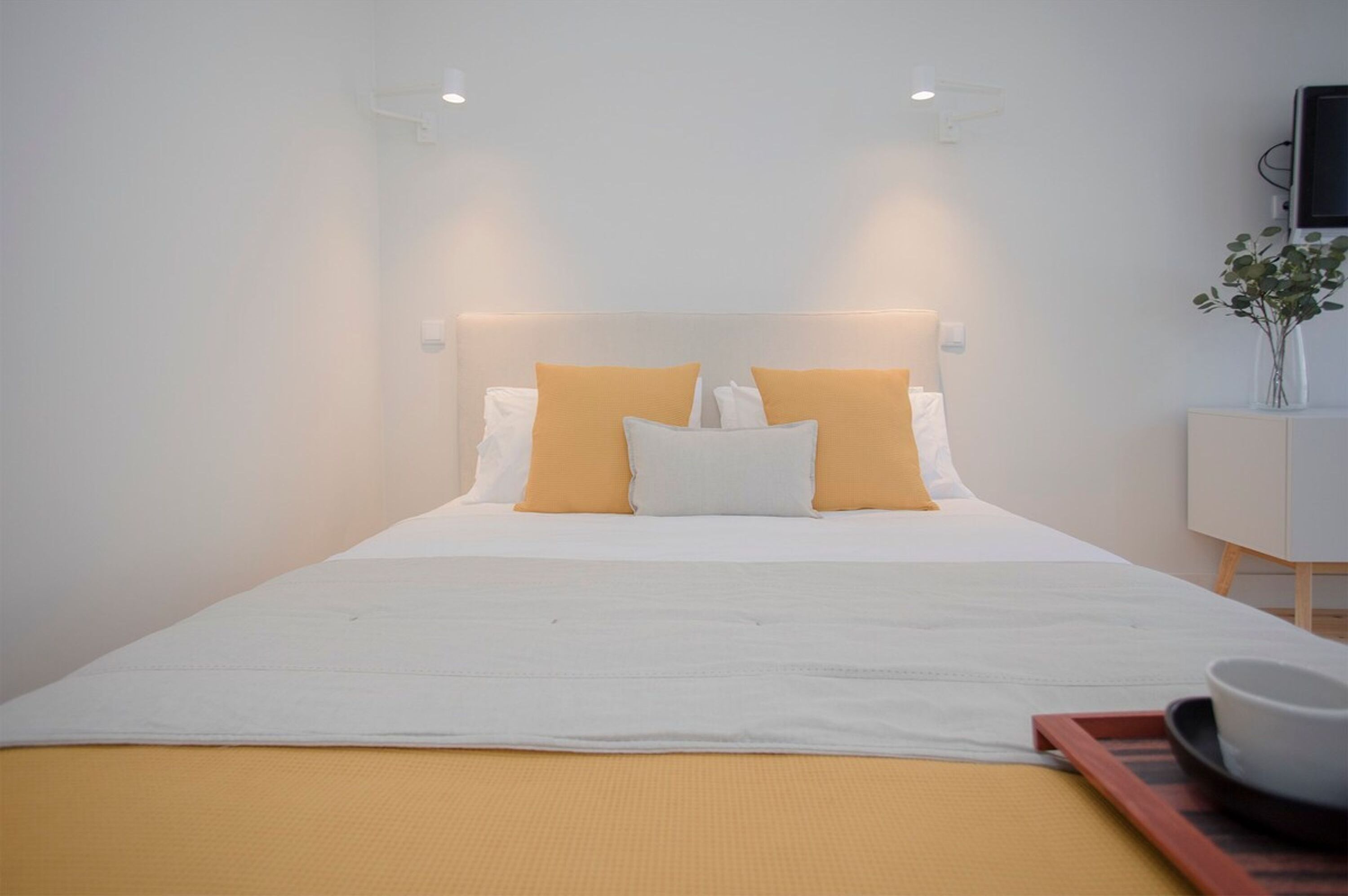 A modern hotel room with a neatly made bed, a bedside table with a vase, and a tray with a bowl and a cup.